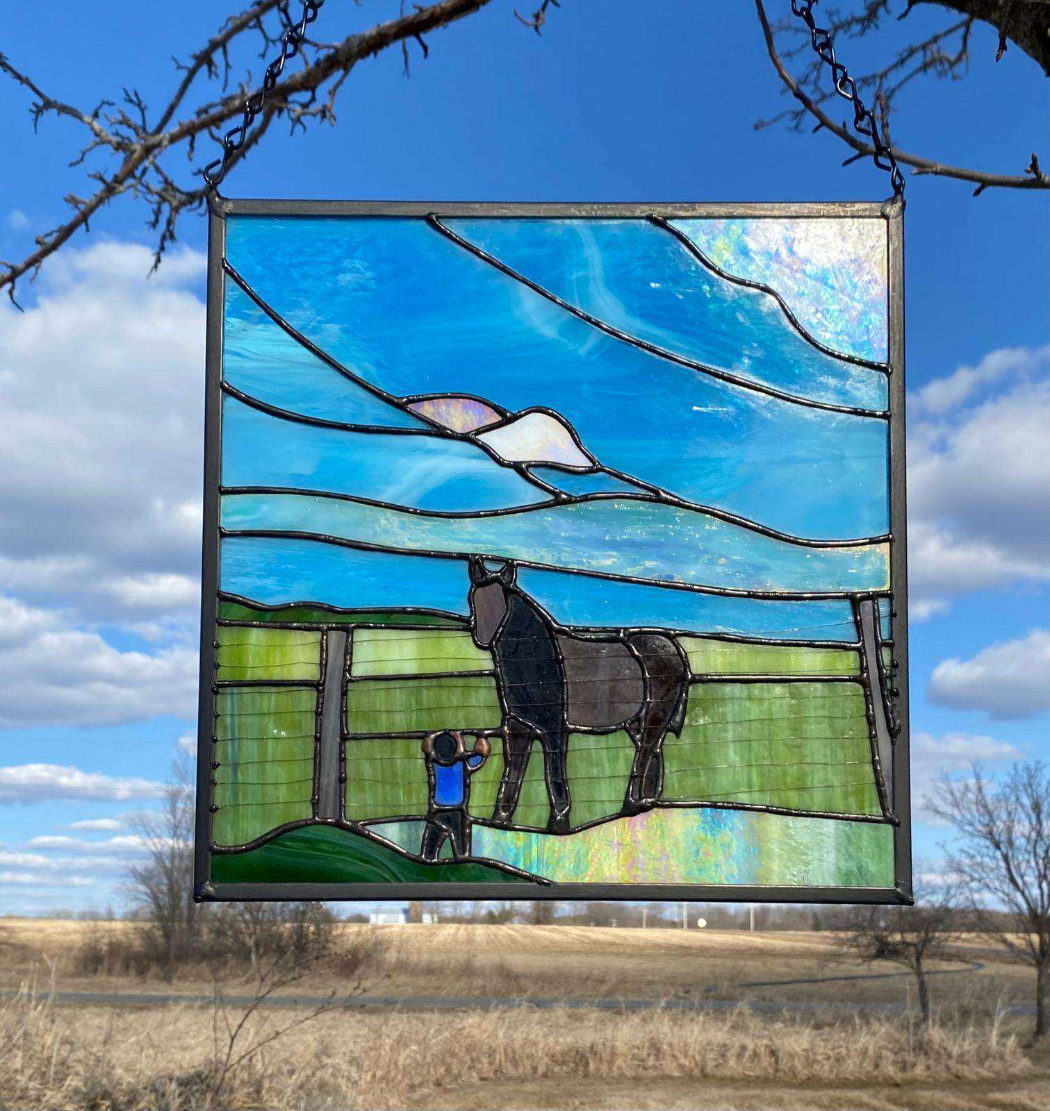 A square stained glass piece of a small boy standing in front of a fence and field with a black and gray horse in it